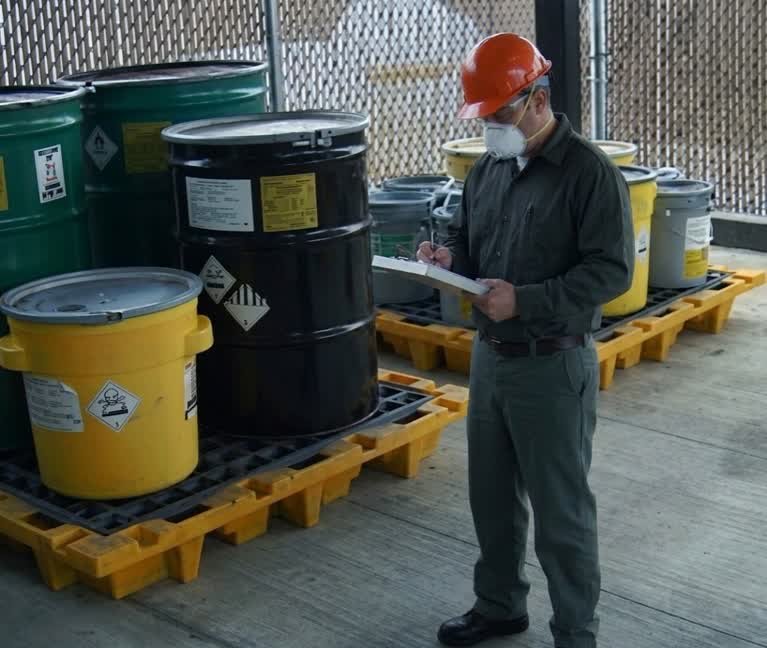 Un inspector con casco naranja y mascarilla realiza el inventario y control de la gestión de residuos industriales en Santa Fe, revisando barriles de sustancias químicas peligrosas.