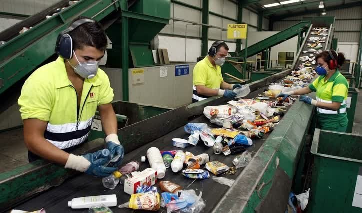 Tres operarios con equipo de bioseguridad realizando la separación y gestión de residuos industriales en Tunjuelito sobre una cinta transportadora en una planta de reciclaje de plásticos.